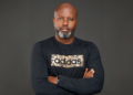 Portrait of Tosh Gitonga, Kenyan filmmaker best known for Nairobi Half Life, standing with arms crossed in a black Adidas shirt against a dark background.