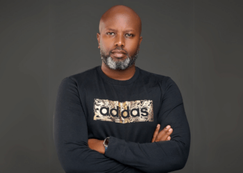 Portrait of Tosh Gitonga, Kenyan filmmaker best known for Nairobi Half Life, standing with arms crossed in a black Adidas shirt against a dark background.