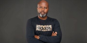 Portrait of Tosh Gitonga, Kenyan filmmaker best known for Nairobi Half Life, standing with arms crossed in a black Adidas shirt against a dark background.